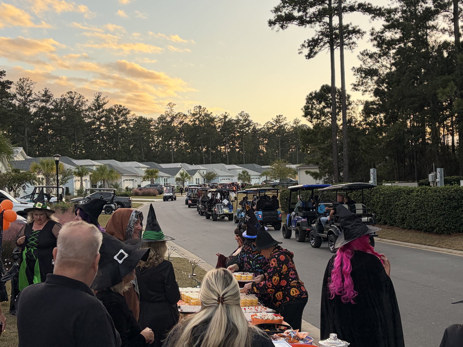 Costumed witches serving food from a roadside table as golf carts wait on the street
